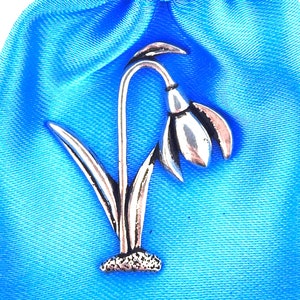 May include: A silver pewter snowdrop flower charm. The flower is white and has a green stem and leaves. The charm is on a blue satin bag.