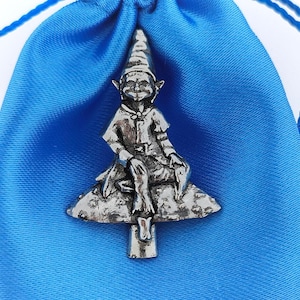May include: A pewter figurine of a gnome sitting on a mushroom. The gnome is wearing a pointed hat and has a long beard. The figurine is a light grey colour and has a detailed design.