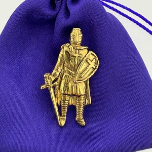 Op de afbeelding: Een gouden tinnen pin met daarop een ridder in harnas met een zwaard en schild. Het schild heeft een kruis erop.