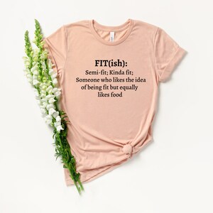 Op de afbeelding: Een lichtroze T-shirt met een zwarte afbeelding die "FIT(ish): Semi-fit; Kinda fit; Someone who likes the idea of being fit but equally likes food." zegt.