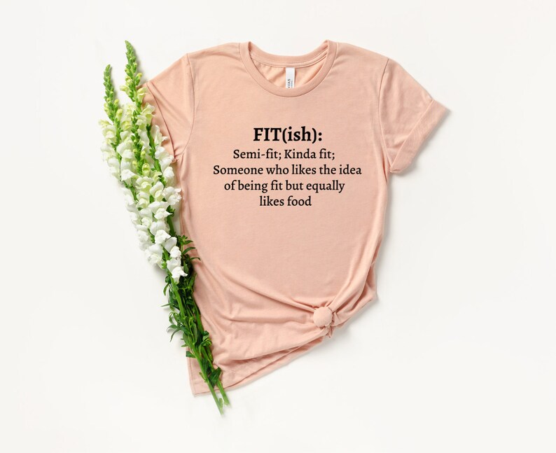 Op de afbeelding: Een lichtroze T-shirt met een zwarte afbeelding die "FIT(ish): Semi-fit; Kinda fit; Someone who likes the idea of being fit but equally likes food." zegt.