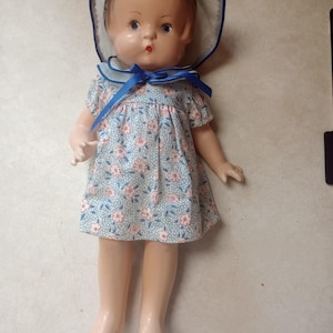 May include: A vintage-style doll with molded brown hair, blue eyes, and a small red mouth. The doll wears a light blue dress with a white and red floral pattern and a matching bonnet featuring a blue ribbon trim and a blue tie under the chin.
