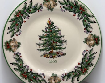 May include: A round, off-white ceramic plate with a green rim and a Christmas theme. The plate features a decorated Christmas tree in the center, surrounded by a garland of pine boughs, red berries, and bells. The year "2005" is printed below the tree.