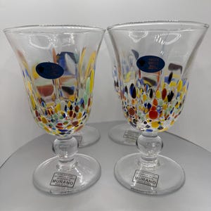May include: Four clear stemmed glasses with colorful speckled designs. The glasses have a rounded bowl shape and a clear stem and base. Each glass has a blue sticker that reads "Vetro Artistico Murano Made in Italy".
