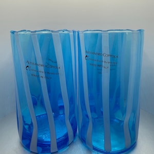 May include: Two cylindrical glass tumblers featuring a vibrant blue hue with vertical white stripes. Each glass displays the black printed text ALESSANDRO COPPOLA Vetreria Arte di Murano 1977 MADE IN ITALY on the front surface of the glass body.