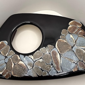 May include: A black ceramic decorative bowl with a unique design. The bowl features a large circular opening and is adorned with a mosaic pattern of gray, brown, and blue stones along the edge. The bowl has a glossy finish.