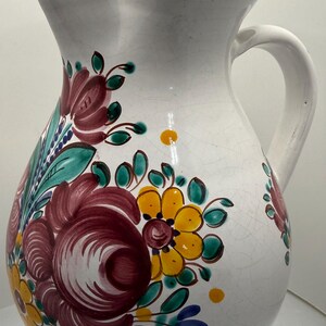 May include: A white ceramic pitcher featuring a hand-painted floral pattern in shades of deep red, yellow, and green. The rim and base are accented with a solid yellow band, and the body displays a subtle crackle glaze finish throughout the surface.