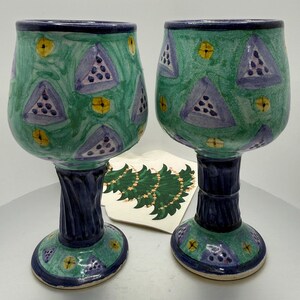 May include: Two hand-painted ceramic wine glasses. The glasses have a green background with blue triangles and yellow accents. The stems are dark blue and twisted. The bases are green with blue and yellow details. A decorative card with a green Christmas tree design is between the glasses.