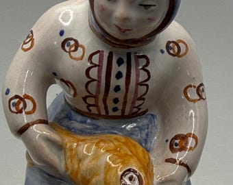 May include: A ceramic figurine of a woman in a blue headscarf and apron, holding a golden fish. The woman's white shirt is decorated with brown pretzel-like shapes and vertical stripes. The figurine has a glossy finish.