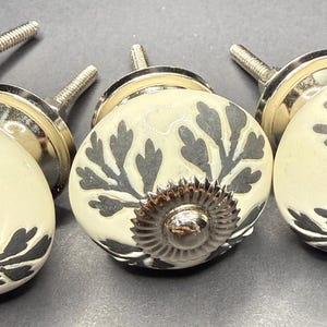May include: Close-up of decorative cabinet knobs. The knobs have a cream-colored base with a dark gray floral design. Each knob has a silver-colored metal base and a silver-colored screw for attachment. The knobs are arranged in a row.