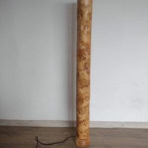May include: A tall, wooden floor lamp with a natural wood finish. The lamp has a cylindrical base and a long, slender shaft. The shaft is covered in a unique wood grain pattern.