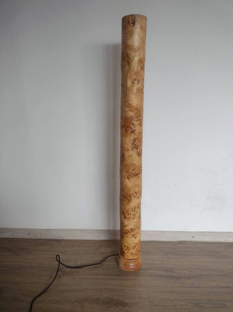 May include: A tall, wooden floor lamp with a natural wood finish. The lamp has a cylindrical base and a long, slender shaft. The shaft is covered in a unique wood grain pattern.
