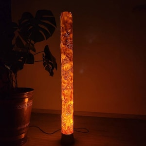May include: A tall, cylindrical floor lamp made of wood with a warm, orange glow. The lamp has a natural wood grain pattern and is lit from within, creating a warm and inviting ambiance.