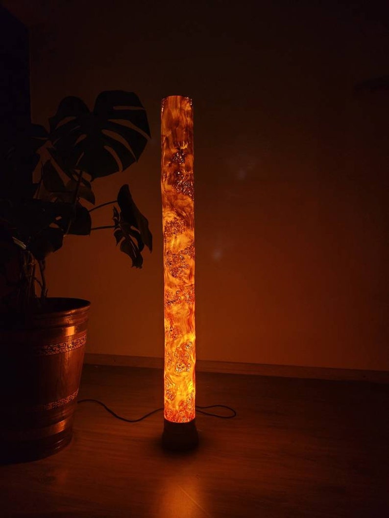 May include: A tall, cylindrical floor lamp made of wood with a warm, orange glow. The lamp has a natural wood grain pattern and is lit from within, creating a warm and inviting ambiance.