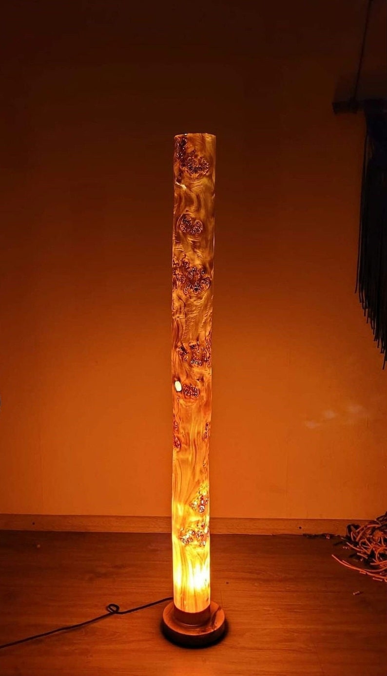 May include: A tall, cylindrical floor lamp made from a natural wood log with a warm, orange glow emanating from the light source inside. The lamp has a simple, wooden base.