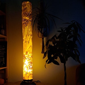 May include: A wooden floor lamp with a cylindrical shade made from a tree trunk. The lamp is lit and casts a warm glow on the surrounding area. A potted plant is visible in the background.