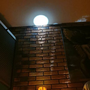 May include: A tall, cylindrical lamp made of wood with a warm, amber glow. The lamp is illuminated from within, casting a soft light on the brick wall behind it. The lamp is in a room with a brick wall and a ceiling with a light fixture.