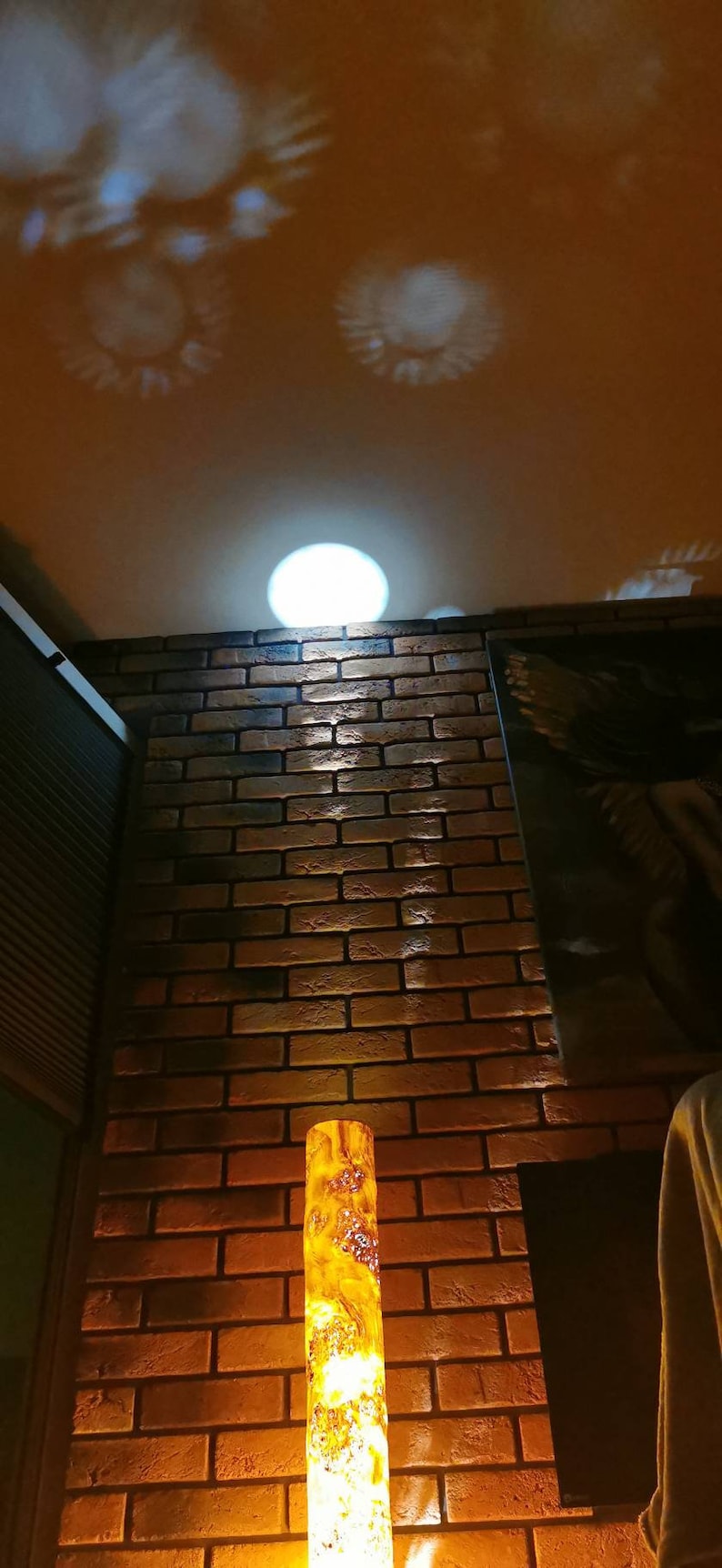 May include: A tall, cylindrical lamp made of wood with a warm, amber glow. The lamp is illuminated from within, casting a soft light on the brick wall behind it. The lamp is in a room with a brick wall and a ceiling with a light fixture.