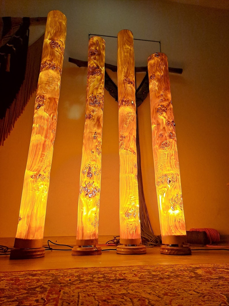 May include: Four tall, cylindrical lamps made from natural wood with a warm, glowing light emanating from within. The lamps are arranged in a row on a wooden floor.