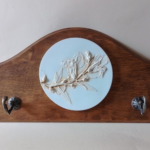May include: Wooden wall-mounted coat rack with a dark brown finish. It features a light blue circular centerpiece with a raised floral design and two decorative metal hooks.