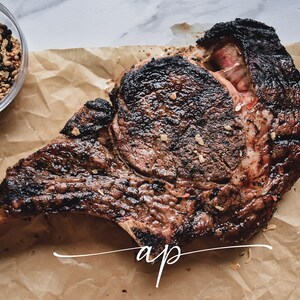 May include: A large, grilled tomahawk steak with a bone, resting on brown butcher paper. The steak is well-done and has a dark brown crust. The steak is seasoned with salt and pepper.