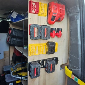 Puede incluir: Un panel de madera con varios soportes para bater&iacute;as de herramientas el&eacute;ctricas y un cargador. Los soportes son rojos, amarillos y negros, y sujetan bater&iacute;as negras y rojas. Un cargador rojo est&aacute; montado en la parte superior.