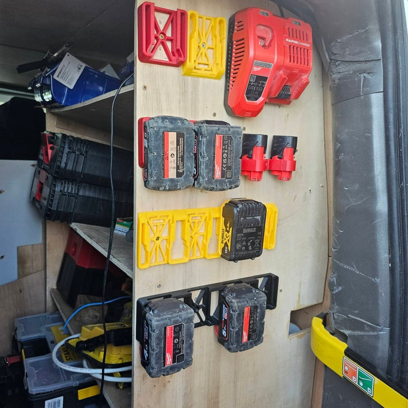 Puede incluir: Un panel de madera con varios soportes para bater&iacute;as de herramientas el&eacute;ctricas y un cargador. Los soportes son rojos, amarillos y negros, y sujetan bater&iacute;as negras y rojas. Un cargador rojo est&aacute; montado en la parte superior.