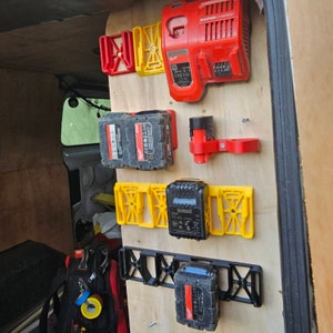 Puede incluir: Una colecci&oacute;n de accesorios para herramientas el&eacute;ctricas, que incluyen soportes de bater&iacute;a rojos y amarillos y un cargador r&aacute;pido rojo, montados en un panel de madera. Tambi&eacute;n se ven varias bater&iacute;as de herramientas el&eacute;ctricas negras y rojas.