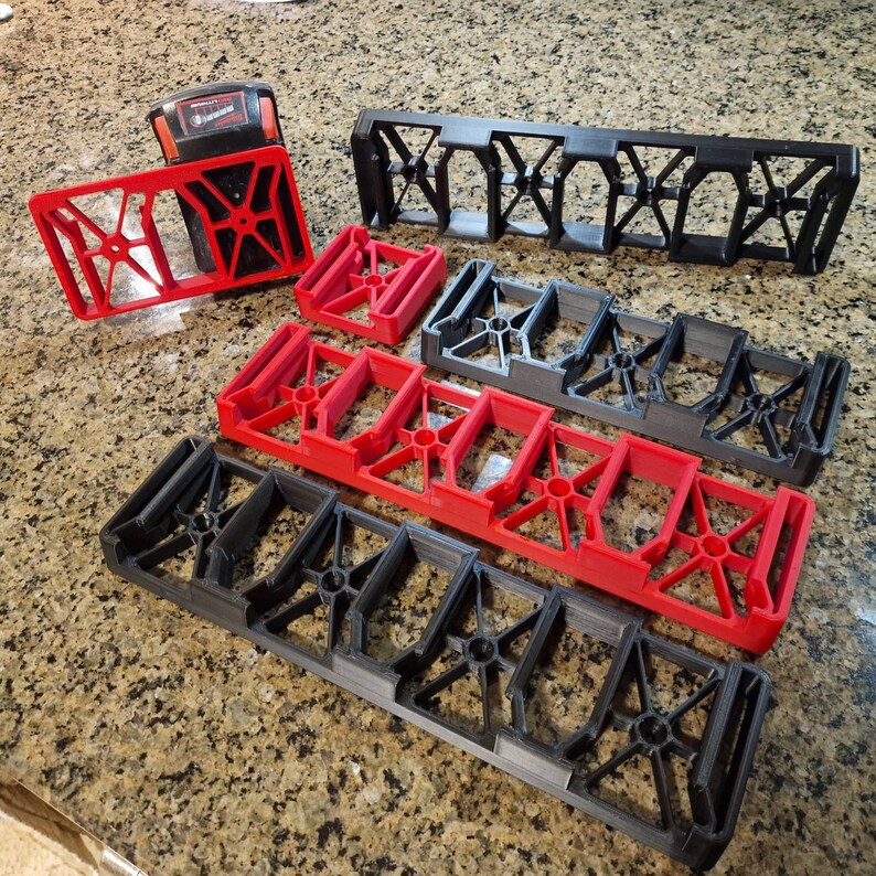 Puede incluir: Una colecci&oacute;n de soportes para bater&iacute;as impresos en 3D, en rojo y negro. Los soportes tienen una estructura geom&eacute;trica de marco abierto y est&aacute;n dispuestos sobre una encimera moteada. Un soporte rojo tiene una bater&iacute;a insertada.