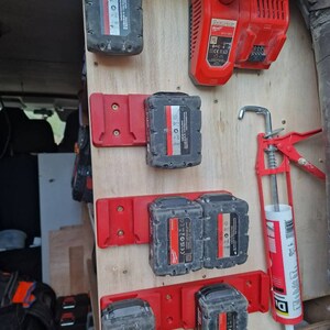 Puede incluir: Un panel de madera con varias bater&iacute;as de herramientas el&eacute;ctricas negras y rojas y un cargador r&aacute;pido rojo. Las bater&iacute;as est&aacute;n montadas en soportes rojos. Tambi&eacute;n se ve una pistola de calafateo roja.