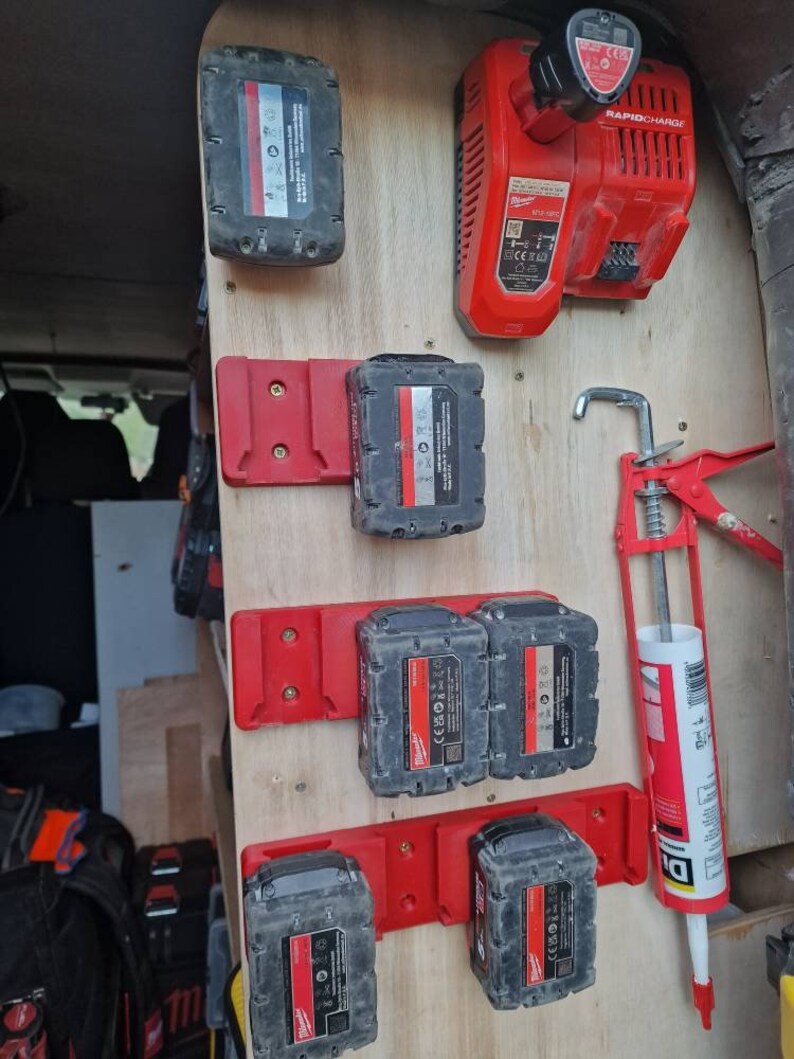 Puede incluir: Un panel de madera con varias bater&iacute;as de herramientas el&eacute;ctricas negras y rojas y un cargador r&aacute;pido rojo. Las bater&iacute;as est&aacute;n montadas en soportes rojos. Tambi&eacute;n se ve una pistola de calafateo roja.