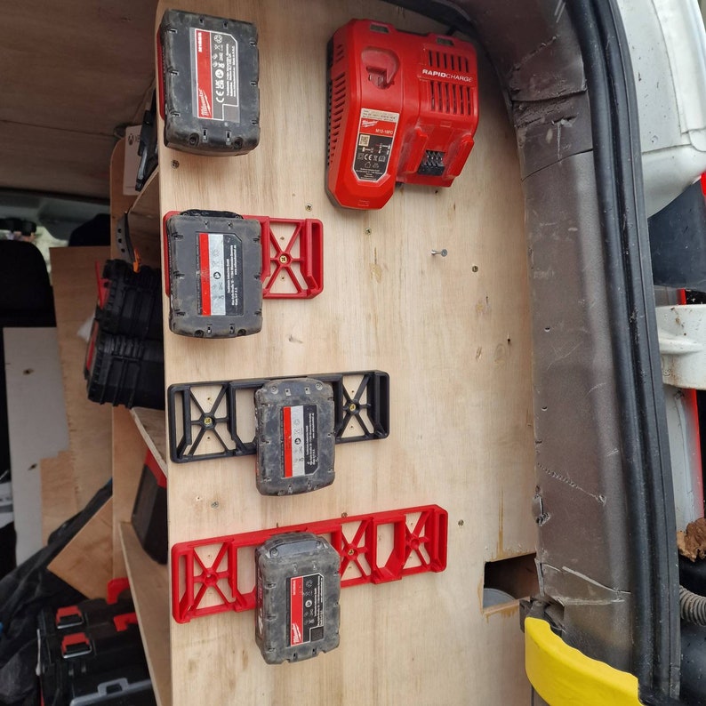 Puede incluir: Un panel de madera dentro de un veh&iacute;culo est&aacute; equipado con soportes rojos y negros para bater&iacute;as de herramientas el&eacute;ctricas y un cargador r&aacute;pido rojo. Varias bater&iacute;as de herramientas el&eacute;ctricas negras y rojas est&aacute;n aseguradas en los soportes. El cargador r&aacute;pido tiene el texto "RAPID CHARGE".