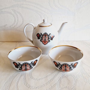 May include: A white teapot and two teacups with a gold rim. The teapot and cups have a black and orange floral design.