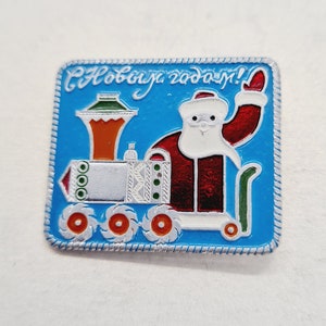 May include: A blue and white enamel pin featuring a cartoon Santa Claus riding a toy train. The train is decorated with red, white and green details. The pin reads "S Novym Godom!" which is Russian for "Happy New Year!"
