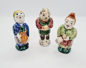 Puede incluir: Tres figuritas de cerámica pintadas a mano que representan personas. La figurita de la izquierda es una mujer que lleva un vestido azul y sostiene una bolsa marrón. La figurita del centro es un hombre que lleva una camisa verde y un chaleco rojo. La figurita de la derecha es una mujer que lleva un vestido verde y sostiene una taza marrón.