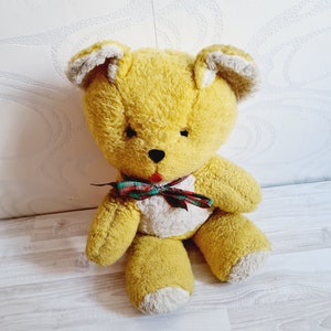 Op de afbeelding: Een gele pluche teddybeer met witte accenten op de oren en poten. De beer draagt een rood-groen geruit strikje.
