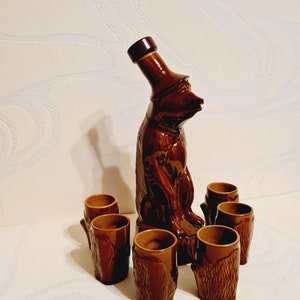 May include: A brown ceramic liquor decanter shaped like a bear with six matching shot glasses. The decanter has a cork stopper and the glasses have a tree bark design.