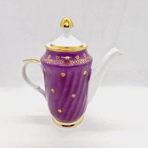 Könnte beinhalten: Eine dekorative lila-weiße Teekanne mit goldenen Akzenten. Die Teekanne hat einen goldverzierten Deckel und Boden, einen weißen Ausguss und Griff sowie ein goldenes Blumenmuster am oberen Rand. Der Korpus der Teekanne hat ein strukturiertes, gewirbeltes Design.