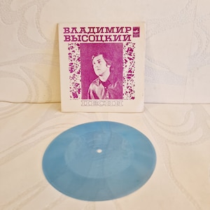 Könnte beinhalten: Eine Vintage-Schallplatte mit einer hellblauen Scheibe und einem rosa Papierhüllen. Die Hülle zeigt ein Schwarzweißfoto eines Mannes mit dem Text "Владимир Высоцкий Песни" in lila.