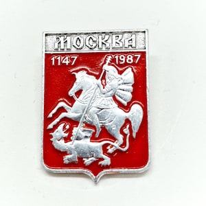 May include: A silver and red enamel pin featuring the coat of arms of Moscow, Russia. The coat of arms depicts a silver knight on horseback slaying a dragon. The pin also includes the text "MOCKBA" and the years "1147" and "1987".