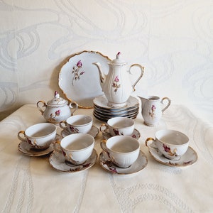 May include: A white porcelain tea set with a rose pattern and gold trim. The set includes a teapot, sugar bowl, creamer, six cups, and six saucers.