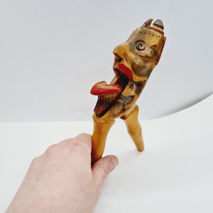 Op de afbeelding: Een houten notenkraker in de vorm van een duivel met een rode tong en beschilderde details. De notenkraker heeft een scharnierende kaak en twee benen.