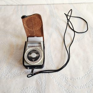 May include: A vintage light meter in a black leather case with a brown leather flap. The meter has a circular dial with numbers and a black strap. The case is open, revealing the meter's face.