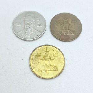 May include: Three South Korean coins: one silver-colored coin featuring a portrait of a man with a beard and hat, and two bronze-colored coins, one darker than the other, both depicting a multi-tiered pagoda structure with Korean text characters.