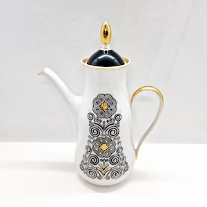 May include: White ceramic coffee pot with a gold lid and handle. The pot has a black and gold geometric design.