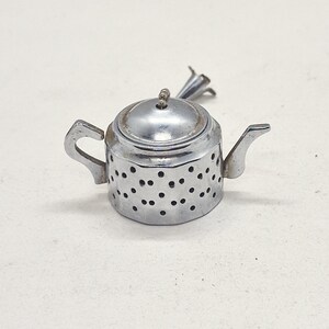 Op de afbeelding: Een kleine, zilverkleurige theepotvormige thee-infuser. De infuser heeft een deksel, een handvat en een tuit. De behuizing van de infuser is geperforeerd met kleine gaatjes. Ontworpen om losse theebladeren te bevatten.