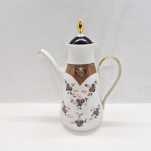 May include: White ceramic coffee pot with a black lid and gold accents. The pot features a brown heart-shaped design with floral patterns and a gold handle. The spout is long and slender.
