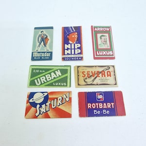 Può includere: Una collezione di sette involucri rettangolari vintage per lamette da barba con vari marchi e illustrazioni, tra cui Matador, Nip Nip, Arrow Luxus, Urban, Severa, Saturn e Rotbart Be-Be con disegni grafici colorati.