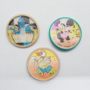 May include: Three round, colorful, cartoon character pins. The first pin features a blue character wearing a crown and standing on a checkered board. The second pin features a black character holding a pear and the text "САМЫЙ БОЛЬШОЙ ДРУГ". The third pin features a yellow character with a red comb and the text "2+2=5".
