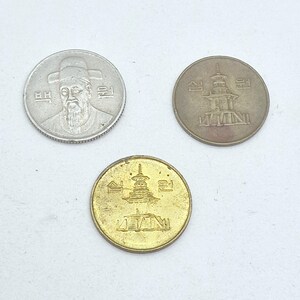 May include: Three South Korean coins: a silver-toned coin featuring a portrait of a man in a traditional hat with the text "백원", and two bronze-toned coins featuring a pagoda illustration with the Korean text "십원" on both sides of the structure.